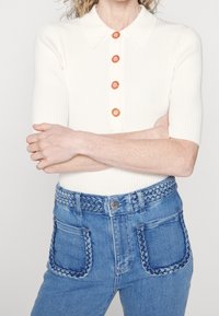 Polo crème à côtes avec des manches courtes et des boutons ronds orange, associé à un jean en denim bleu avec des accents de poches tressées.