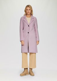 Lila wollen jas met een geforceerde kraag, twee zwarte knopen en lange mouwen, gecombineerd met beige wijde pantalons en tan veterschoenen.