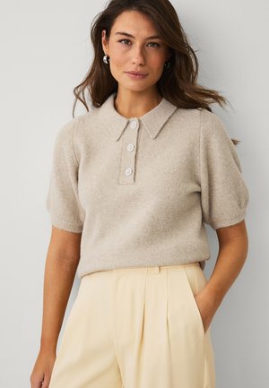 Kvinde med langt brunt hår iført en beige strikket top med korte ærmer, krave og tre knapper, kombineret med lyse cremegule højttaljede bukser.