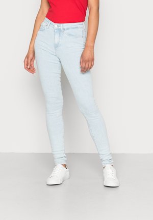 Person iført lyseblå skinny jeans, rød top og hvide sneakers står mod en ensfarvet lys baggrund.