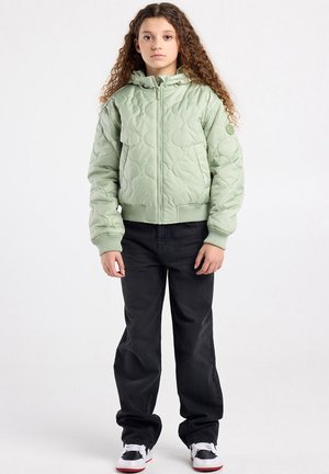 Jeune personne aux cheveux bouclés portant une veste matelassée vert clair, un pantalon ample noir et des baskets blanches, debout devant un fond uni.