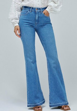 Vrouw draagt lichtblauwe high-waist flared jeans met een witte blouse met bloemenprint, bruine sandalen met open teen en één hand in de zak.