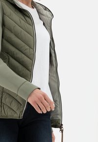 Giacca imbottita verde oliva con zip e design trapuntato, interno grigio e polsini a coste, indossata sopra una maglietta bianca.
