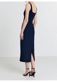Robe midi sans manches bleu marine avec fente latérale, coupe ajustée, tissu lisse et fermeture éclair dans le dos. Talons noirs à brides visibles.