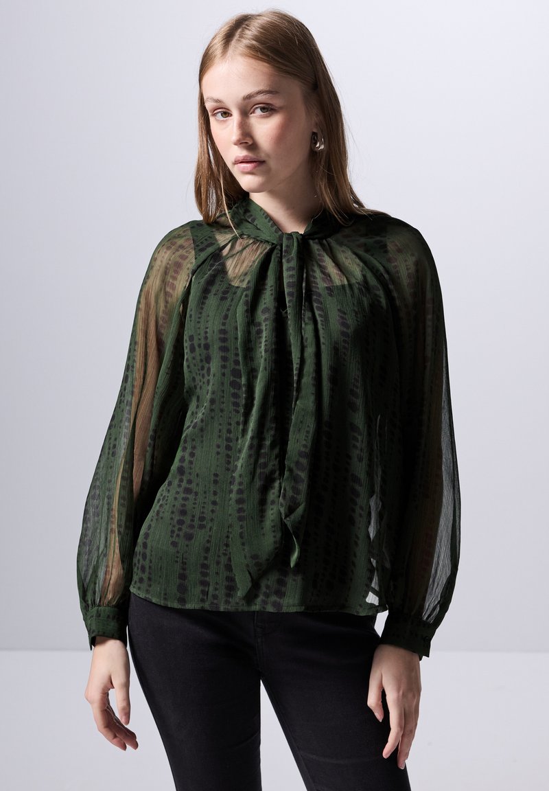 Blusa verde trasparente con maniche lunghe, scollo a fiocco e motivo a righe verticali di un verde più scuro. Abbinata a pantaloni neri.