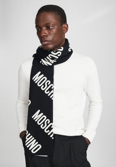 Sort strikket tørklæde med fremtrædende hvid "MOSCHINO" tekst i et gentaget mønster. Blød tekstur, draperende stil, båret over en hvid sweater.