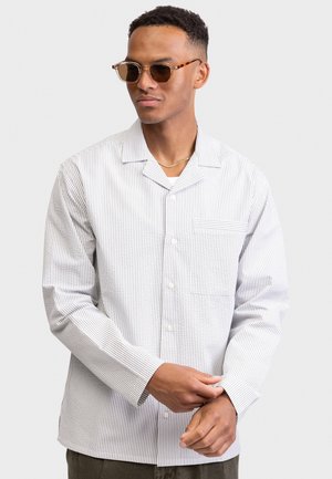 Mand iført skildpaddemønstrede solbriller, guldkæde og hvid langærmet skjorte med tynde lodrette striber, der justerer manchet.