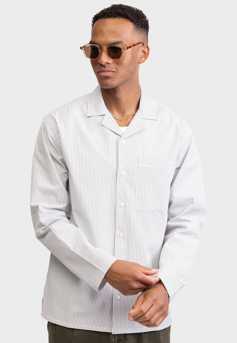 Άνδρας που φορά γυαλιά ηλίου με μοτίβο χελώνας, χρυσή αλυσίδα και λευκό πουκάμισο με μακριά μανίκια και λεπτές κάθετες ρίγες, ρυθμίζει το μανίκι του.