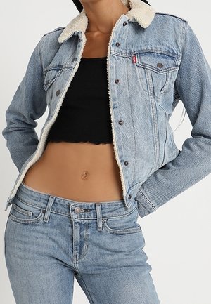 Vrouw die een lichtblauwe spijkerjas draagt met witte fleece voering, een zwarte cropped top en bijpassende lichtblauwe spijkerbroek, staand met handen in de zij.