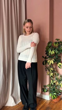 Ung kvinde iført hvid kabelstrikket sweater og sorte bukser, justerer ærmet og står ved siden af en grøn potteplante foran en lyserød væg.