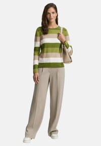 Grün- und cremefarbener gestreifter Strickpullover, kombiniert mit beigen weit geschnittenen Hosen und weißen Sneakers. Das Model trägt eine beiges Handtasche.