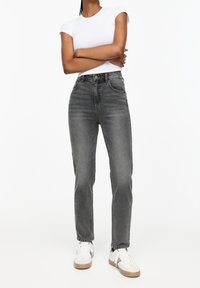 Jean taille haute à jambes droites en denim gris délavé. Comprend des poches avant, une fermeture par boutons, et des ourlets cousus. Porté avec des baskets blanches.