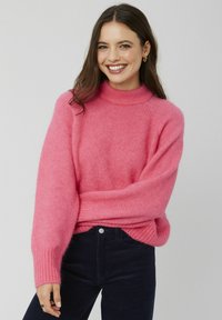Un pull en tricot rose vif avec un col haut et côtelé. Il présente des manches longues amples et une texture douce. Associé à un pantalon en velours côtelé foncé.