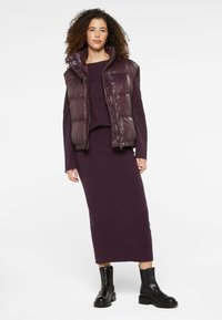 Pflaumenfarbene Pufferweste mit hohem Kragen, über einem abgestimmten langärmligen Kleid. Schwarze Stiefeletten runden das Outfit ab. Glatte und strukturierte Oberflächen.