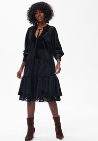 Czarna, trzywarstwowa sukienka o miękkiej fakturze, z dekoltem w kształcie litery V i zawiązywaniem, zdobiona haftowaną koronką na dole oraz długimi rękawami.