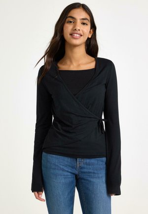 Nuori nainen, jolla on musta kietaisutyylinen pitkähihainen paita ja siniset farkut, seisoo vaalean valkoisen taustan edessä.