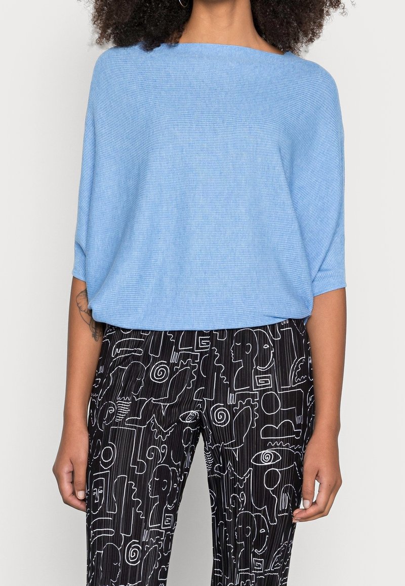 Hellblauer, gerippter Pullover mit einem Bootsausschnitt, kombiniert mit schwarzen Hosen, die weiße abstrakte Linienzeichnungen und vertikale Streifen aufweisen.