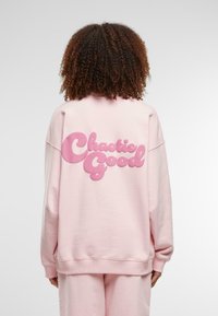 Rosa hettegenser med overdimensjonert "Chaotic Good"-tekst i en dristig, boblede design på baksiden. Myk stoff med avslappet passform og ribbestrikkede ermer.