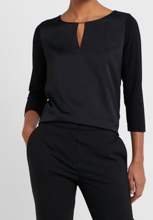 Persoon draagt een zwart shirt met lange mouwen en een kleine split bij de halslijn aan de voorkant en zwarte broek, staand met één hand in de zak.
