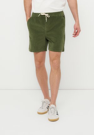 Mand iført olivengrønne shorts med snøre, hvide og sorte sneakers samt en hvid skjorte, stående mod en ensfarvet lys baggrund.