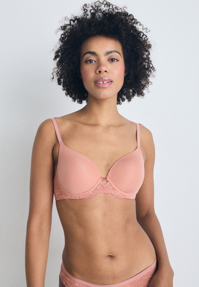 Reggiseno rosa chiaro con coppe lisce e bordi in pizzo; presenta spalline regolabili e un dettaglio decorativo in metallo al centro.