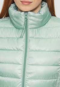 Hellgrüne Puffjacke mit hohem Kragen, durchgehendem Reißverschluss und gestepptem Muster. Das Material hat einen sanften Glanz und verfügt über Kordelzüge.