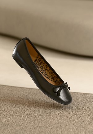 Ballerine en cuir noir avec un petit nœud sur le dessus et une doublure intérieure imprimé léopard reposant sur une surface beige texturée.
