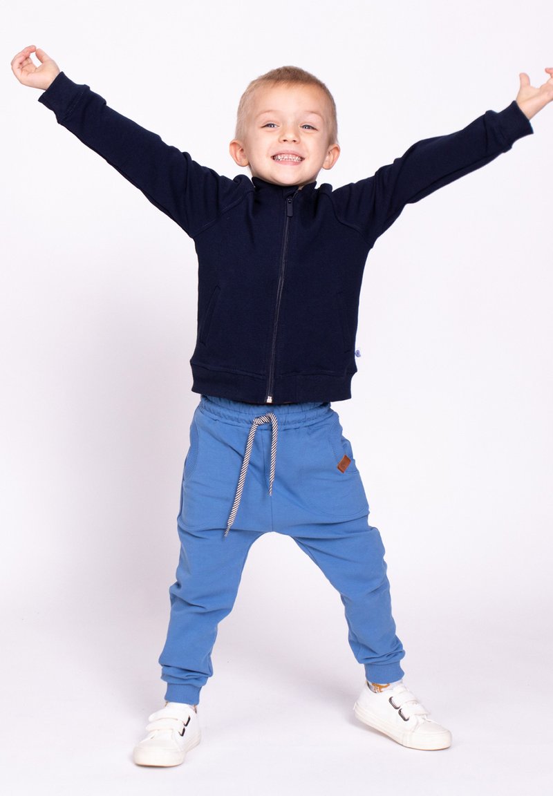 Granatowa bluza z zamkiem i wysokim kołnierzem, zestawiona z niebieskimi dresami z troczkami, o teksturowanym wykończeniu i boczną kieszenią. Całość dopełniają białe trampki.