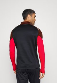 Schwarzes langärmliges Sportshirt mit roten Ärmeln, einem strukturierten braunen Kragen, körperbetonter Passform und glattem Stoff. Keine Grafiken oder Muster sichtbar.