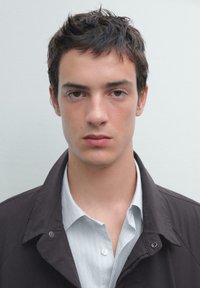 Giacca nera sopra una camicia a righe blu chiaro. Primo piano di un giovane uomo con capelli castani corti e espressione neutra. Illuminazione soffusa.