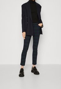 Person klädd i svart polotröja, marinblå dubbelknäppt kavaj, grön och marinblå rutiga slim-fit byxor och svarta kraftiga loafers på en enfärgad bakgrund.