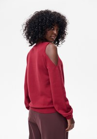 Kobieta z kręconymi włosami, ubrana w czerwony sweter z odsłoniętymi ramionami i brązowe spodnie, spoglądająca przez ramię na tle białego tła.