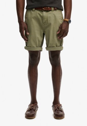 Hombre con pantalones cortos oliva enrollados, cinturón trenzado marrón, reloj de cuero marrón y zapatos casuales marrones con cordones, de pie con las manos relajadas.