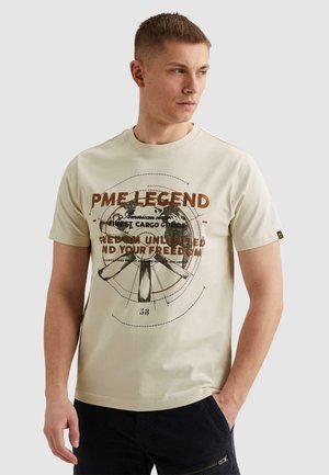 Hombre vistiendo una camiseta de manga corta beige con texto gráfico y diseño de globo, combinada con pantalones negros, posando frente a un fondo liso.