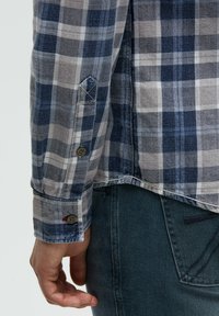 Karierte Hemd in Blau- und Grautönen, mit Knopfabnäher, sichtbaren Nähdetails und einem leicht abgerundeten Saum. Über einer dunklen Jeans getragen.