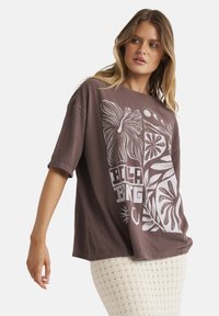 Frau trägt ein übergroßes braunes T-Shirt mit grafischen Blumen- und Mondmotiven, kombiniert mit einem cremefarbenen, strukturierten Rock, vor einem weißen Hintergrund.
