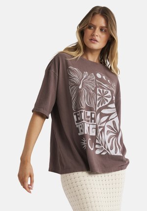 Woman wearing oversized brown graphic t-shirt with floral and moon designs, paired with a cream textured skirt, against a white background.