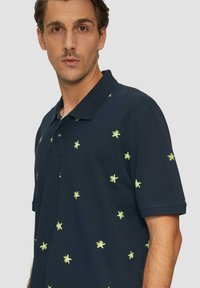 Granatowa koszulka polo z krótkim rękawem, zdobiona rozproszoną haftowaną dekoracją w jasnym zielonym kolorze, z klasycznym kołnierzykiem i zapięciem na guziki.