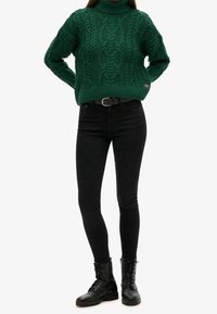 Vihreä kaapeli-neule turtleneck-paita paksulla tekstuurilla, yhdistettynä mustiin istuviin farkkuihin ja mustiin nauhakuosisin taistelubootseihin.