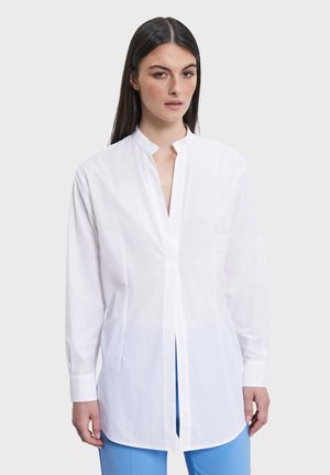 Vrouw met lang donker haar, gekleed in een wit tuniekshirt met lange mouwen en een blauwe broek, staand tegen een effen lichte achtergrond.