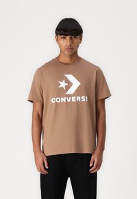 Ruskea puuvillapaita, jossa on valkoinen tähti, nuoli ja "CONVERSE" -painatus. Lyhythihaisena ja pyöreällä kaula-aukolla.