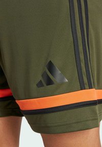Oliwkowe szorty sportowe z czarnymi paskami, pomarańczowymi akcentami na dolnym brzegu oraz czarnym logo Adidas w dolnym lewym rogu.