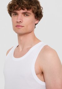 Vit bomullstopp utan ärmar med rundad hals och breda armhål, som visar en slät textur och minimala sömmar. Modell med kort, krulligt hår.