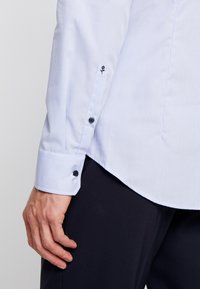 Vaalean sininen ruudullinen pitkähihaine paita, jossa on mustat napit ja pieni musta brodeerattu ruusu hihassa.