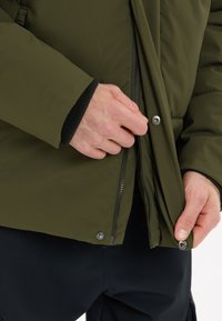 Olijfgroene geïsoleerde jack met een ritssluiting aan de voorkant, drukknoopsluiting en zwarte ribgebreide manchetten. Handen die de ritssluiting en het knoopgedeelte aanpassen.
