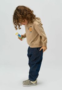 Hanorac bej cu un grafic în formă de stea, pantaloni fracți cu potrivire lejeră din denim închis, și adidași slip-on în carouri alb-negru. Copil care ține un tub.