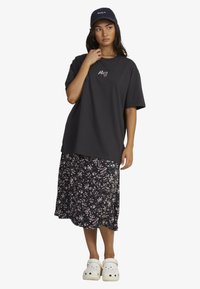 Zwarte oversized t-shirt met geborduurd logo, gecombineerd met een midi-jurk in bloemmotief. Witte klompen maken de outfit compleet.