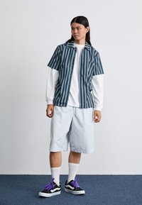 Camicetta a righe a maniche corte in teal e bianco, sovrapposta a una maglietta bianca a maniche lunghe. Pantaloni corti azzurri e scarpe da ginnastica nere con lacci viola.