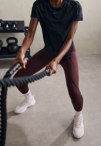 Czarny sportowy T-shirt, bordowe legginsy z ozdobnymi szwami oraz jasnoróżowe buty sportowe. Trzyma gruby, czarny worek treningowy na siłowni.