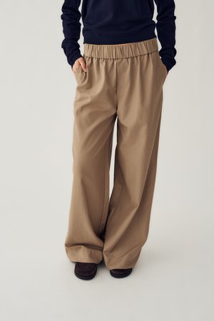 Osoba w luźnych beżowych spodniach z elastycznym pasem i bocznymi kieszeniami, ubrana w ciemny top i ciemne buty, stojąca na jasnej podłodze.